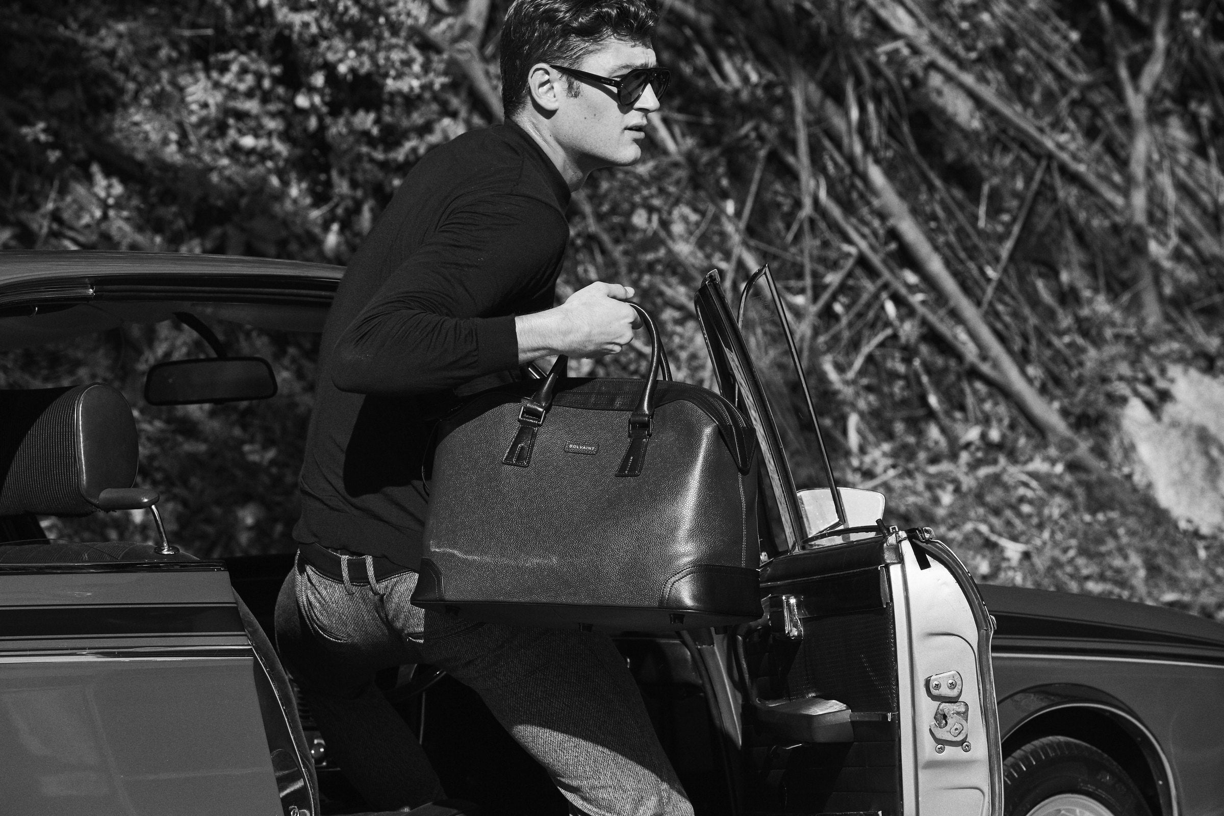A man lifting a black leather travel bag into a vintage convertible