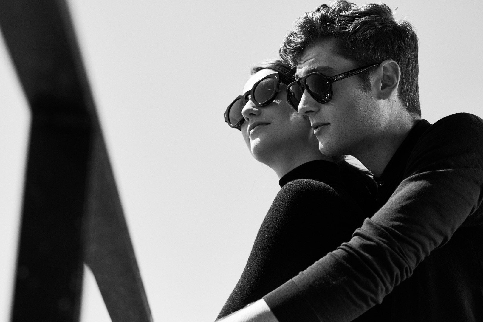 A couple in black, both in dark sunglasses, looking out against a bright sky